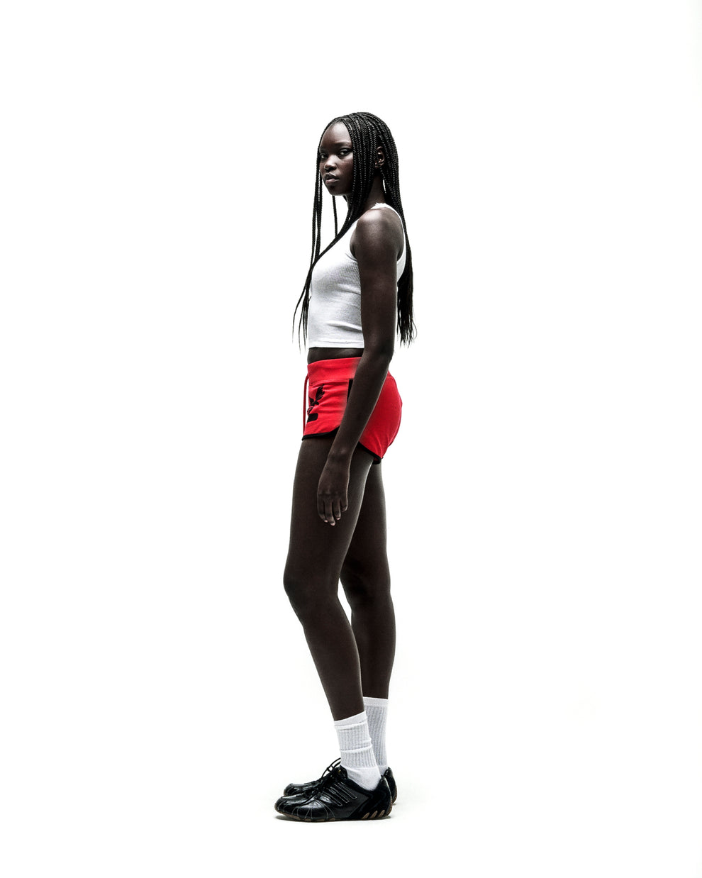 Woman in SP5 WIDE BOXER red shorts with black logo, white tank top, and black sneakers.